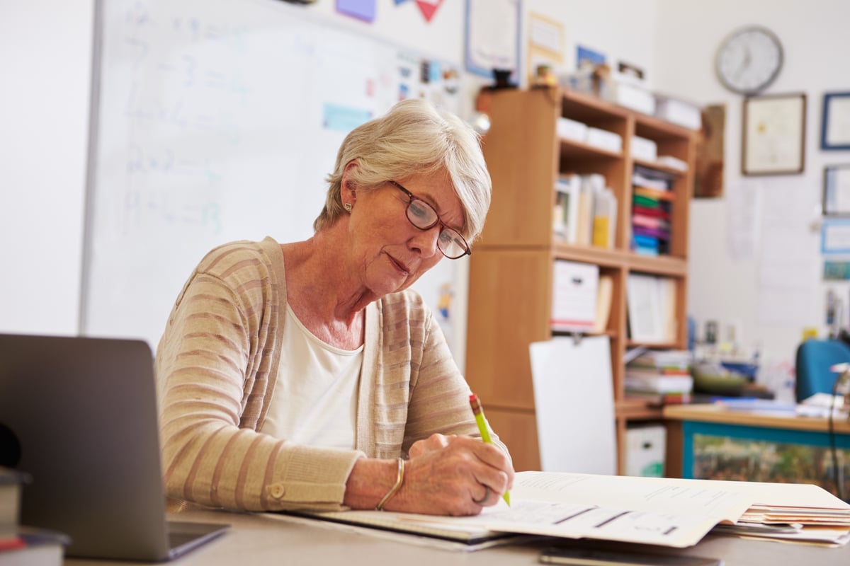 A senior teacher sits at her classroom desk marking.