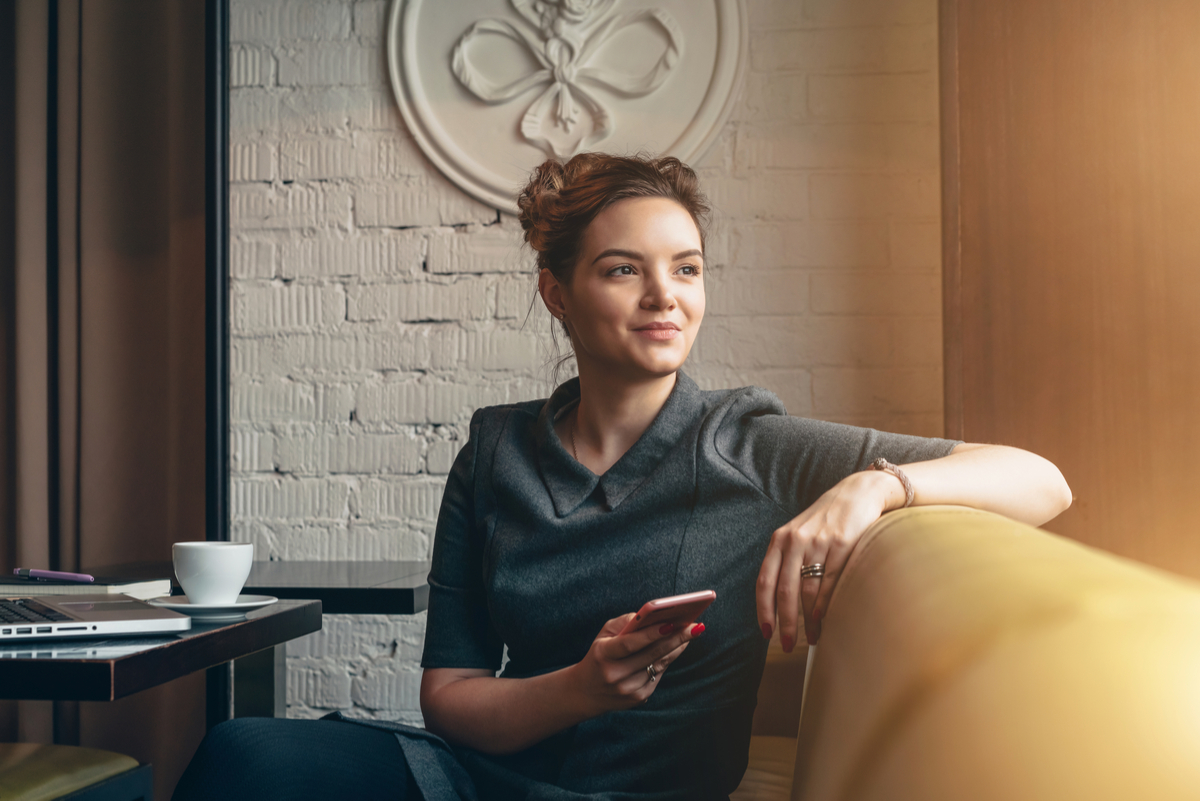 A young, smiling woman sits in a cafe holding her phone.