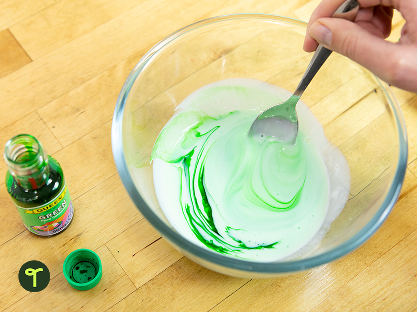 A teacher's hand stirs green food colouring into a bowl of slime on a desk in the classroom