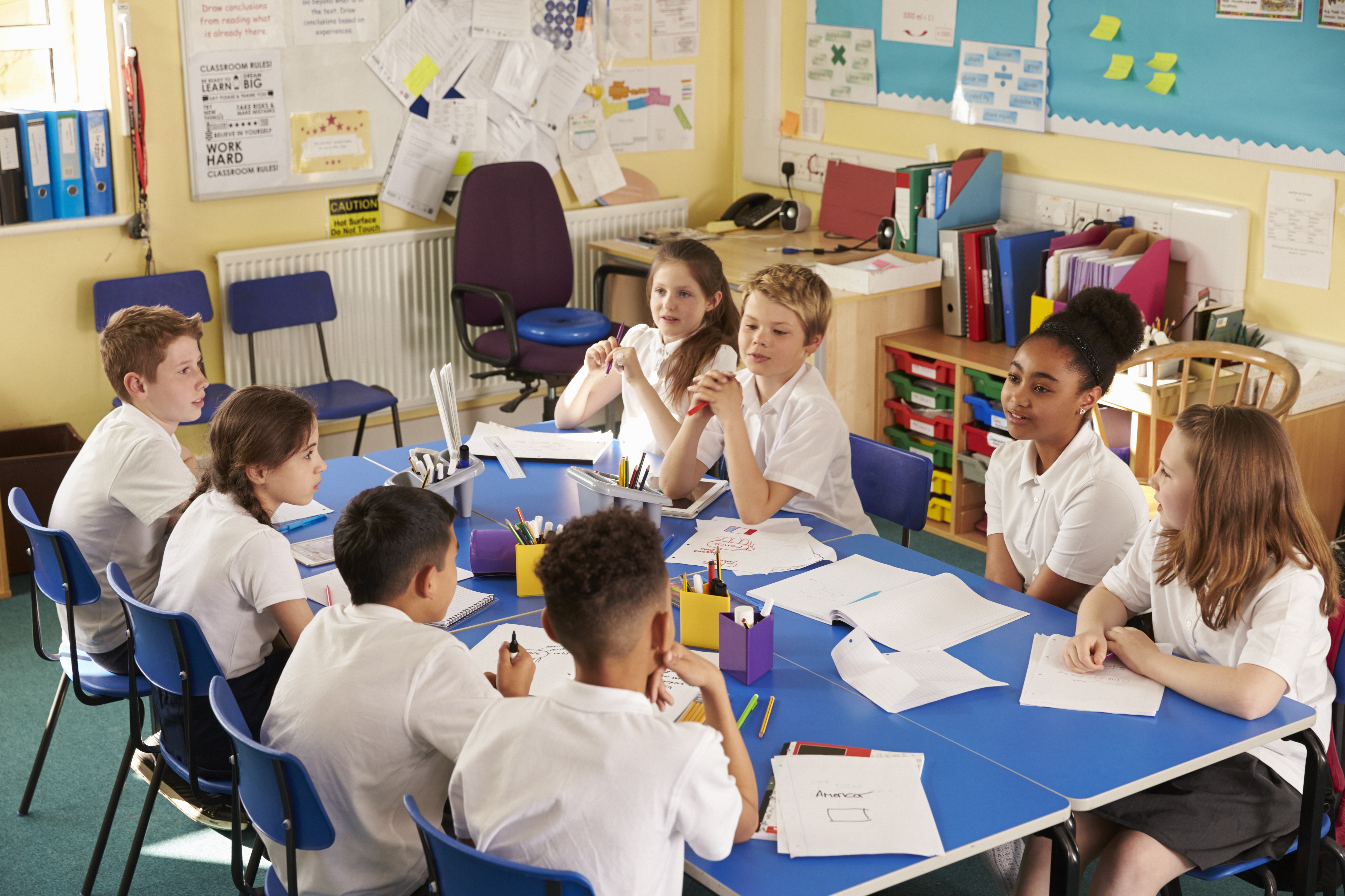  A group of students sitting around a table in a classroom having a discussion with their teacher.