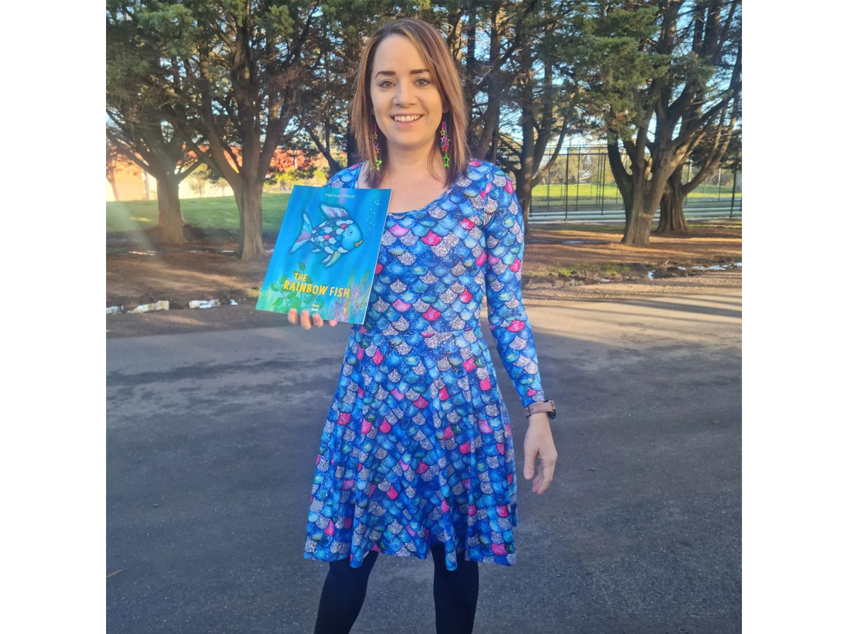 A woman wearing a sparkly blue and purple dress and holding the Rainbow Fish book
